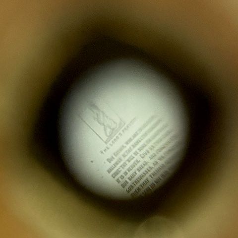 Looking through one of the Stanhope viewers showing the micro Lord's Prayers. It also depicts a child on their knees saying their prayers. Collection of the author.